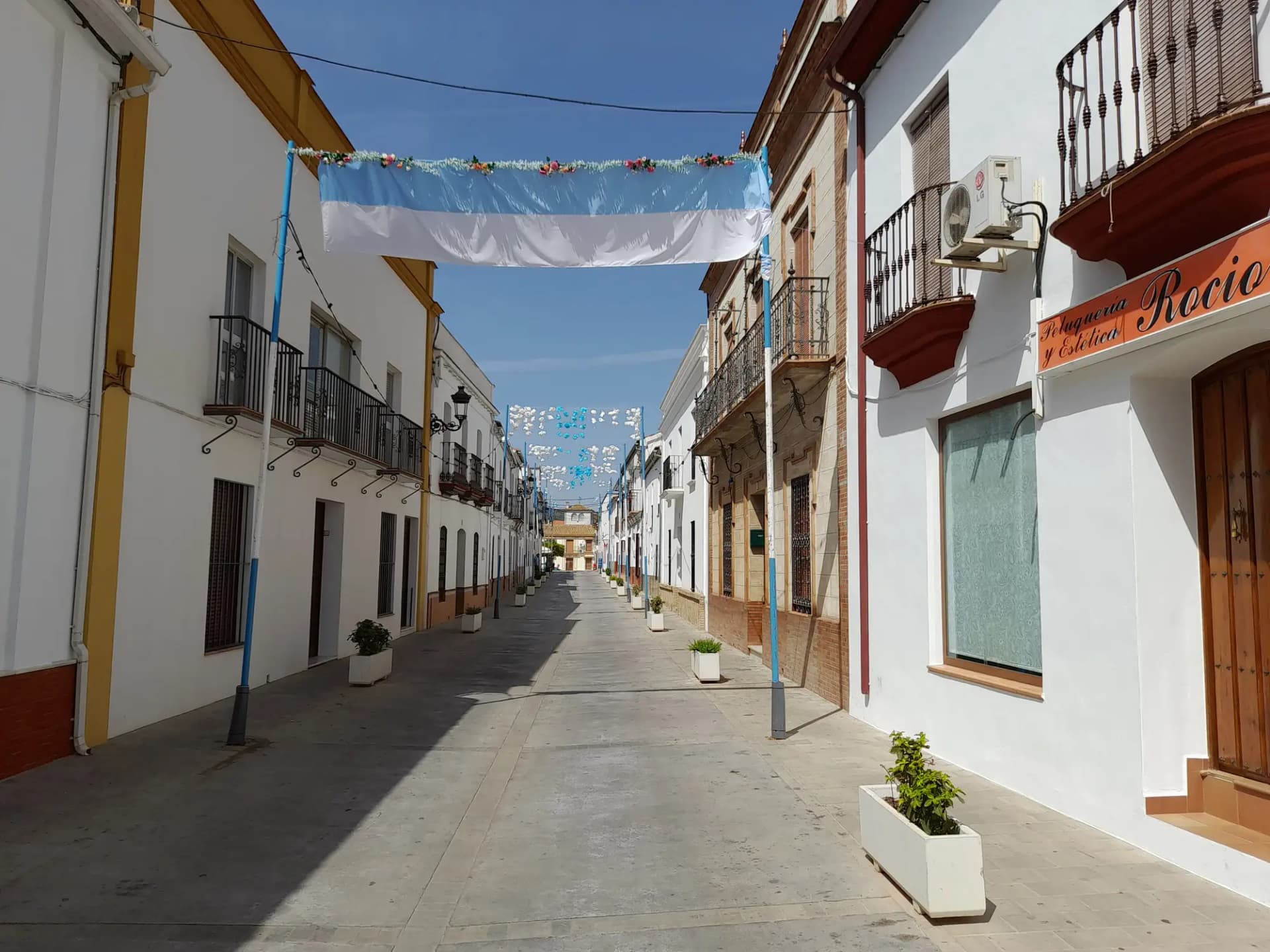 Calle de casas blancas con balcones y un toldo de fiesta colgado entre fachadas en Las Navas de la Concepción
