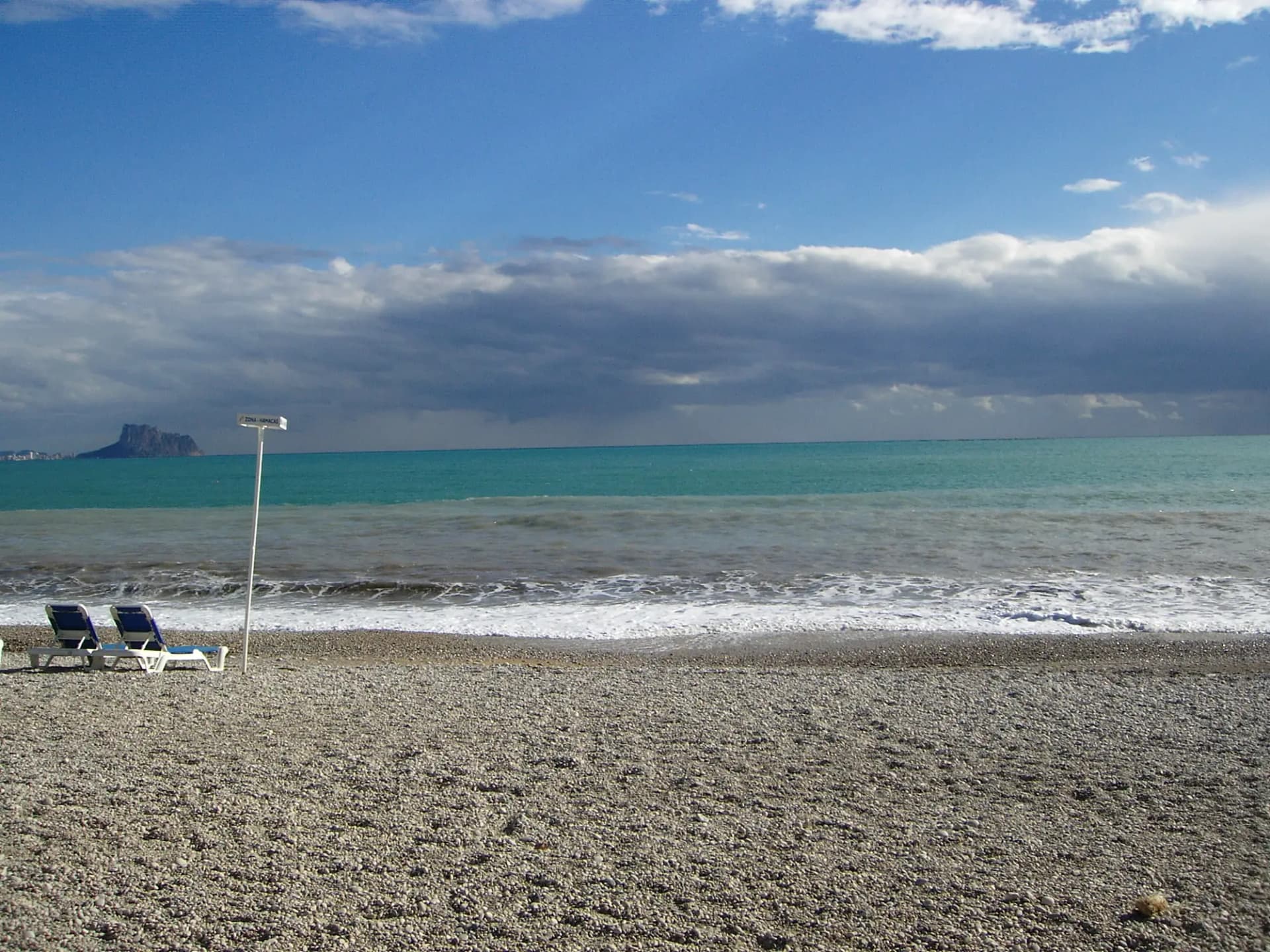 Playa de cantos del Racó del Albir con el mar en calma y nubes sobre el horizonte