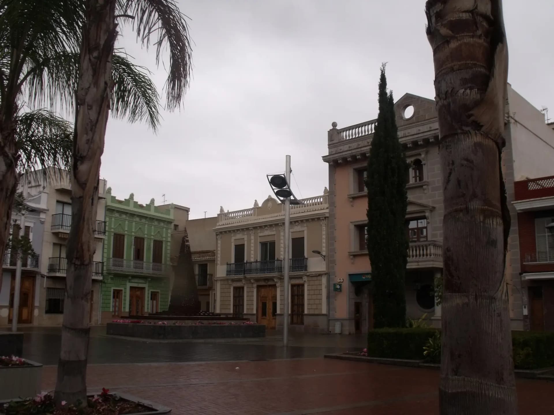 Plaza de Picassent con edificios de principios del siglo XX, palmeras y un ciprés en una tarde de lluvia