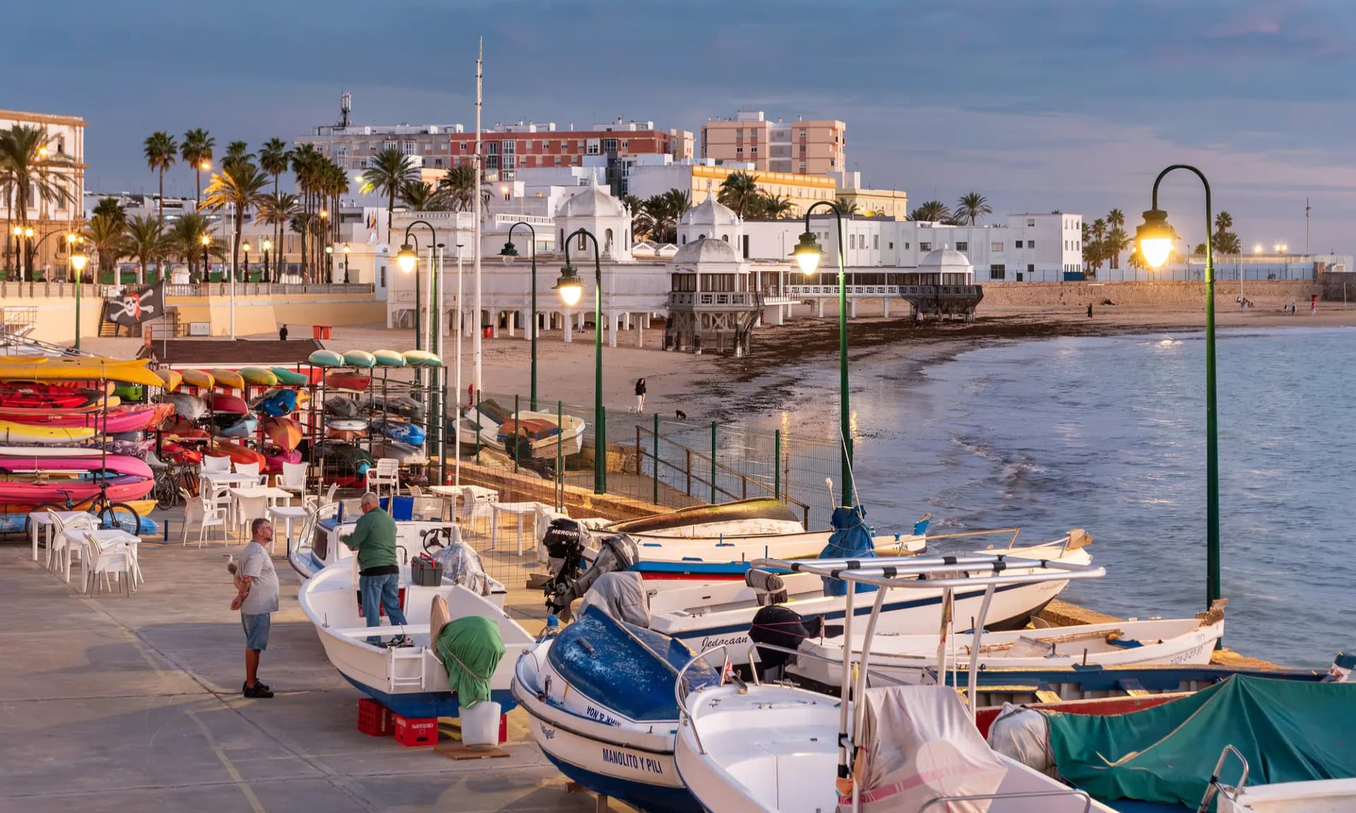 Playa de La Caleta con barcas varadas, paseo con farolas y casas blancas al atardecer