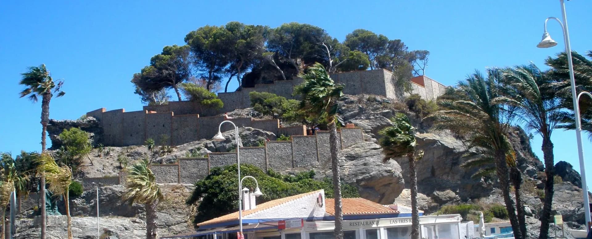 Peñón del Santo de Almuñécar con muros de piedra, pinos y palmeras junto al paseo marítimo