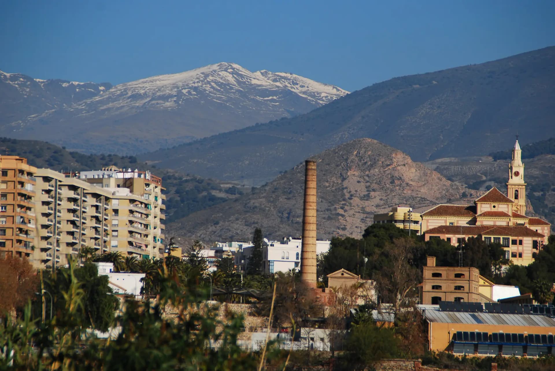 Edificios de Motril con una alta chimenea de ladrillo y una torre de iglesia, y las cumbres nevadas de Sierra Nevada al fondo
