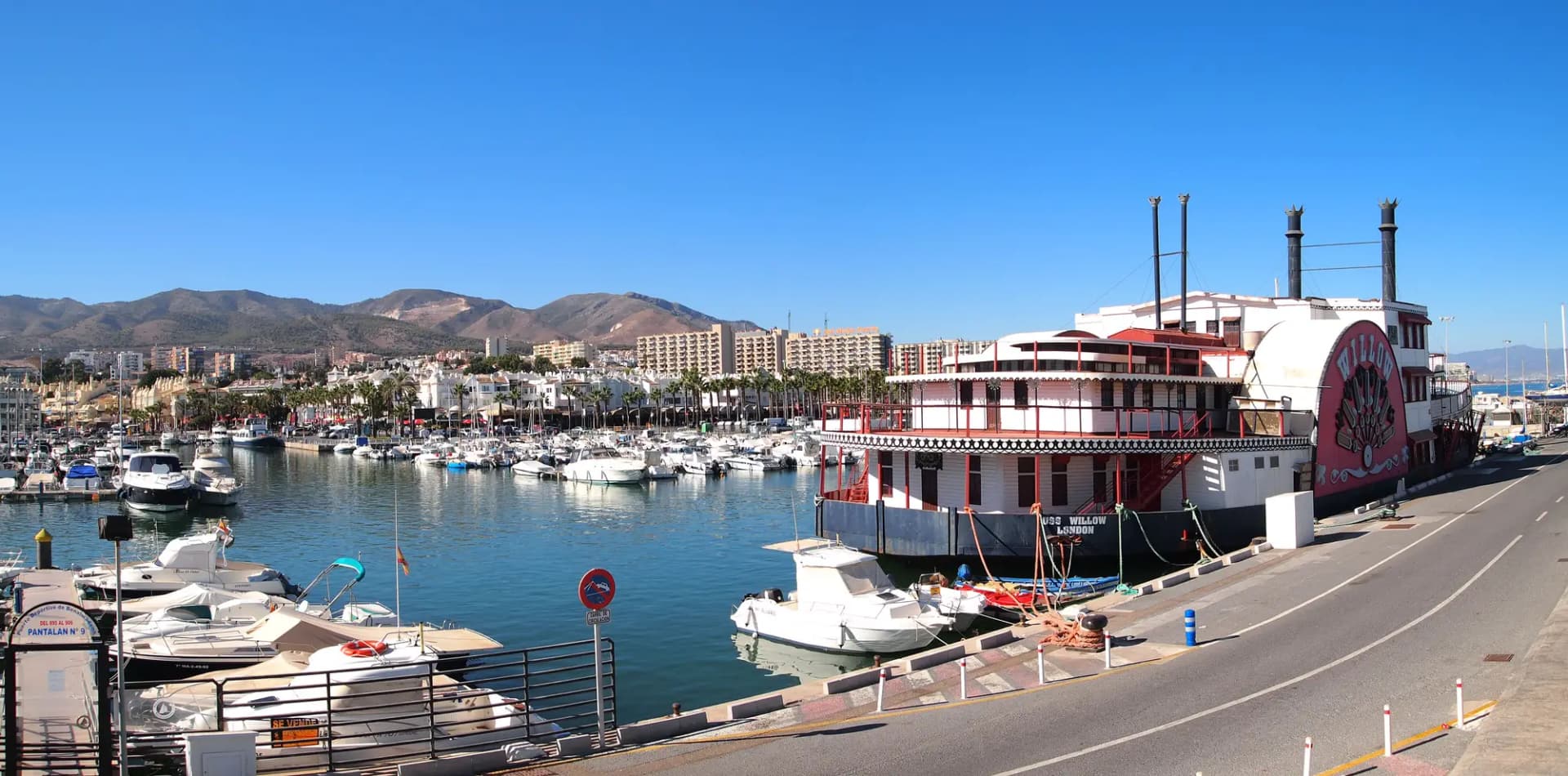 Puerto deportivo con barcos amarrados, un barco de ruedas de paletas y edificios blancos con la sierra al fondo