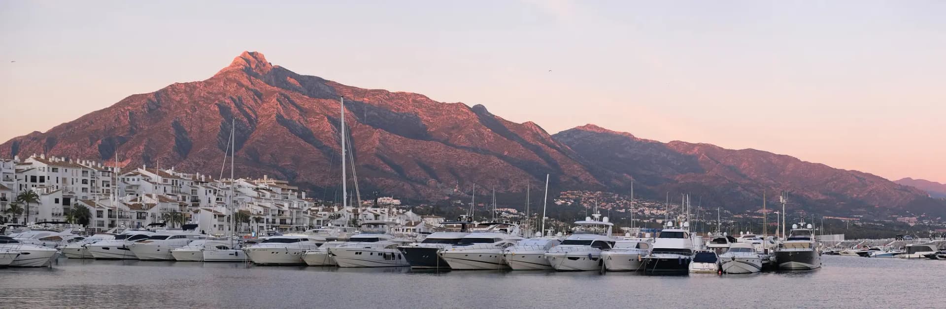 Yates amarrados en Puerto Banús con edificios blancos y la sierra al fondo al atardecer