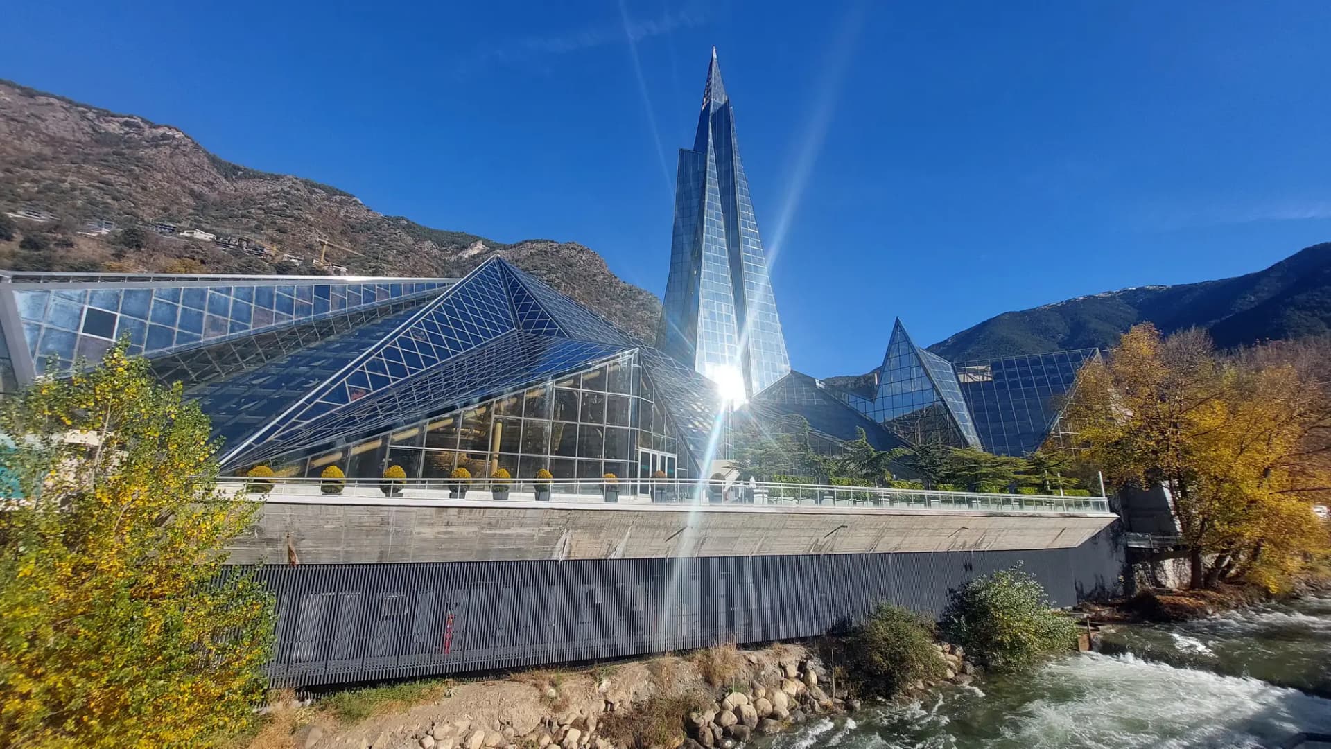 Edificio de cristal de Caldea, con su aguja puntiaguda, junto al río Valira y con montañas al fondo