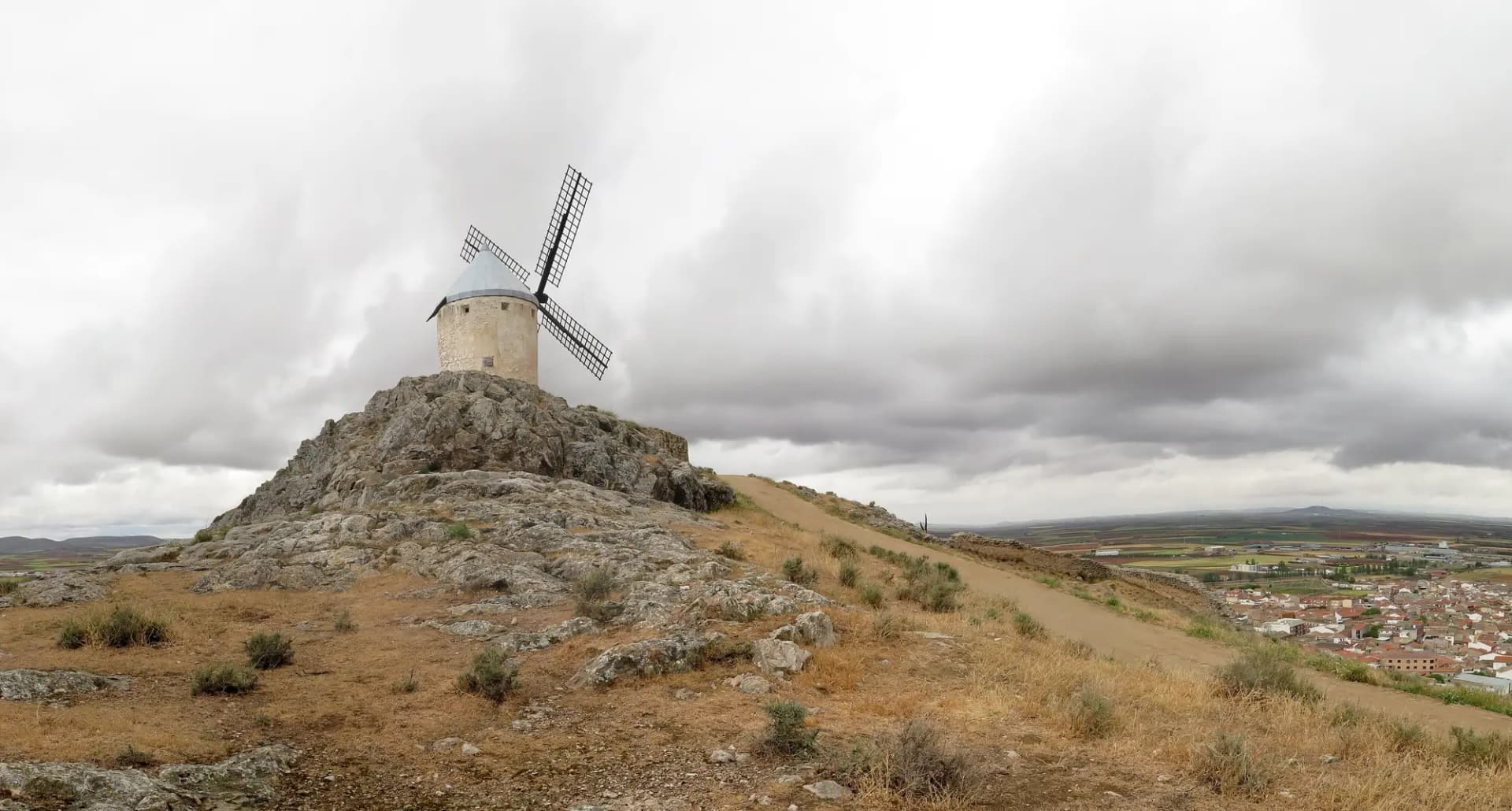 Molino de viento blanco sobre un cerro rocoso con el pueblo de Consuegra y la llanura al fondo