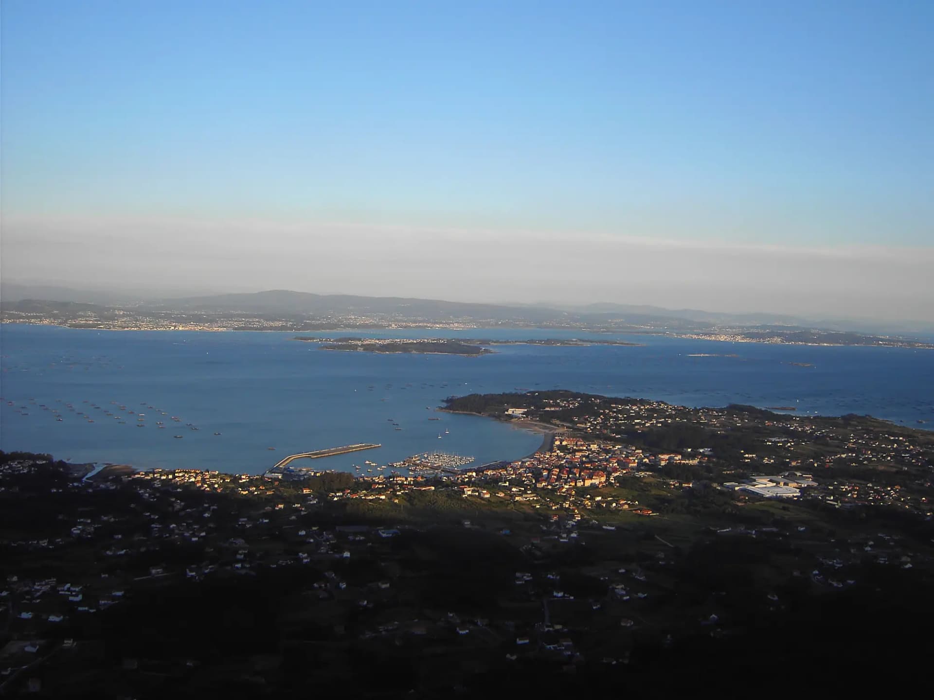 Ría de Arousa vista desde lo alto, con bateas de mejillón en el agua, un puerto y la costa al fondo