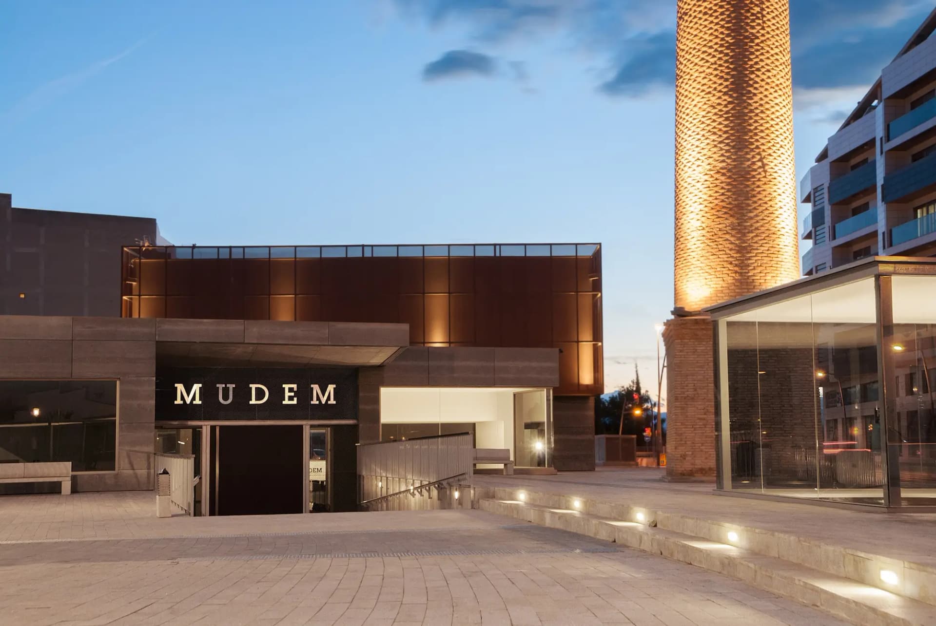 Edificio del MUDEM y antigua chimenea industrial iluminados al anochecer en Molina de Segura